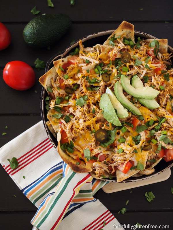 A skillet full of chicken nachos, topped with salsa, cheese, peppers, and sliced avocado.