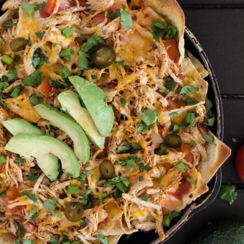 A skillet full of chicken nachos, topped with salsa, cheese, peppers, and sliced avocado.